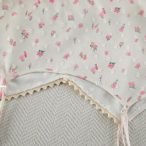 Puede incluir: Primer plano de una prenda blanca con un delicado estampado floral de rosas rosas y hojas verdes. La prenda tiene un ribete de encaje festoneado en color crema. La tela parece ligera y aireada.