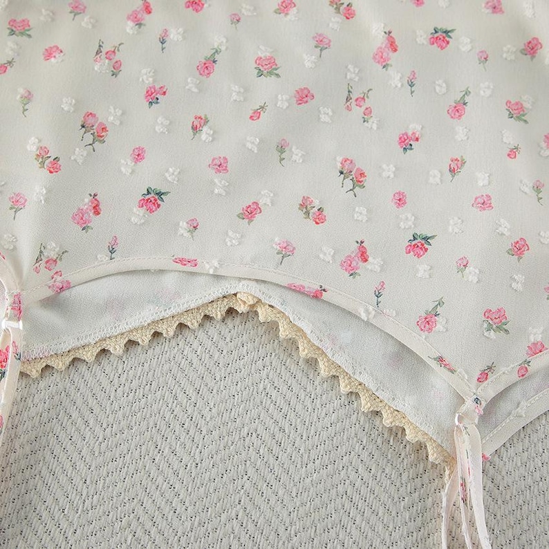 Puede incluir: Primer plano de una prenda blanca con un delicado estampado floral de rosas rosas y hojas verdes. La prenda tiene un ribete de encaje festoneado en color crema. La tela parece ligera y aireada.