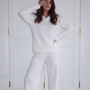 以下が含まれることがあります： 襟付きのVネック長袖プルオーバーと、お揃いのワイドレッグパンツで構成された、無地の白いツーピースラウンジウェアセット。滑らかで不透明な生地で作られており、すっきりとしたモノクロームの美しさを特徴としています。