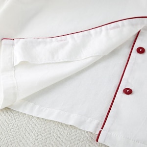 May include: Close-up of a white garment with red trim and buttons. The fabric is folded, revealing the inside lining. The red trim outlines the edges and button placket, with two visible red buttons.