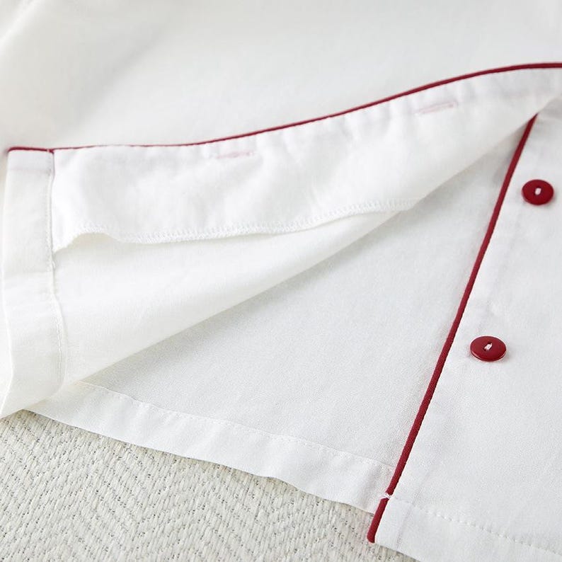 May include: Close-up of a white garment with red trim and buttons. The fabric is folded, revealing the inside lining. The red trim outlines the edges and button placket, with two visible red buttons.
