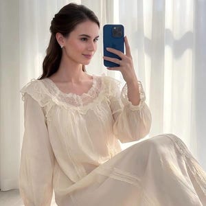 Pode incluir: Uma camisola de cor creme de manga comprida com decote com folhos, acabamentos em renda delicada no colarinho e nos punhos, e painéis de renda verticais na parte da frente. A peça é feita de um tecido leve e semi-transparente.