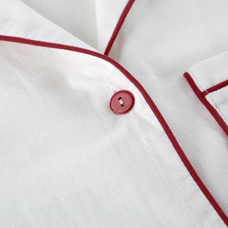 May include: Close-up of white pajama top with red trim and a matching red button. The fabric appears to be a lightweight material, possibly linen or cotton. The red trim outlines the collar and button placket.