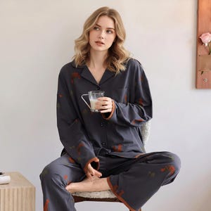 May include: Dark gray pajama set with a button-down shirt and pants. The set features a pattern of orange and green shapes and brown trim. The model is holding a clear glass mug with a white liquid.