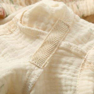 May include: Close-up of a cream-colored fabric with a textured, crinkled appearance. A beige herringbone-patterned strap is attached to the fabric with visible stitching. The fabric appears to be part of a garment, possibly a top or dress.