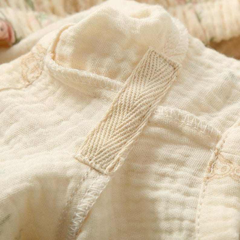 May include: Close-up of a cream-colored fabric with a textured, crinkled appearance. A beige herringbone-patterned strap is attached to the fabric with visible stitching. The fabric appears to be part of a garment, possibly a top or dress.