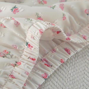 Puede incluir: Primer plano de una tela color crema con un delicado estampado floral. La tela presenta peque&ntilde;os puntos en relieve y est&aacute; adornada con motivos de rosas rosas y rojas. Los bordes est&aacute;n fruncidos, creando un efecto de volantes, y la textura general parece suave y ligera.