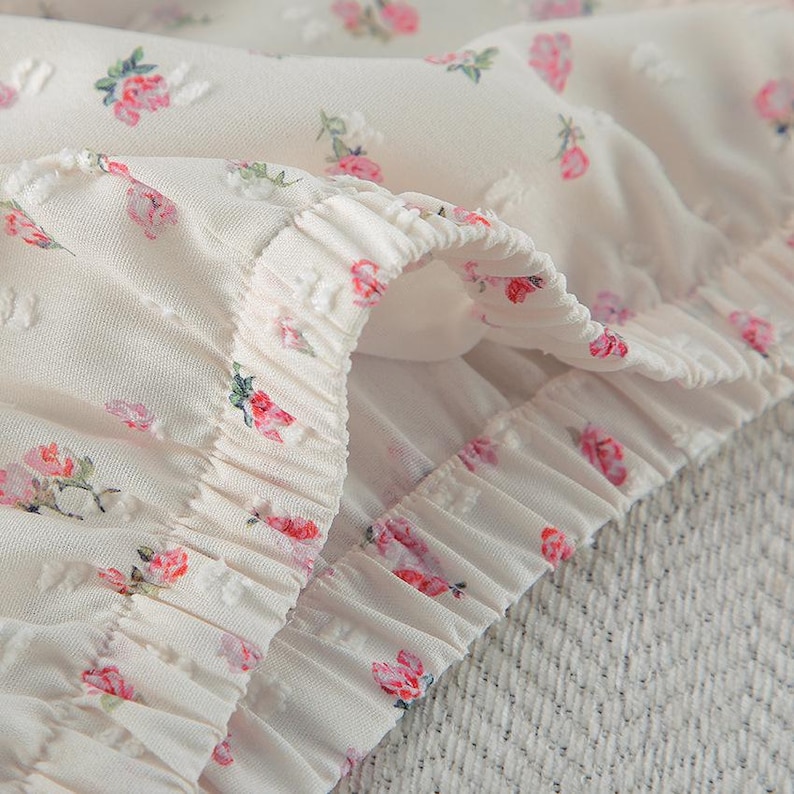 Puede incluir: Primer plano de una tela color crema con un delicado estampado floral. La tela presenta peque&ntilde;os puntos en relieve y est&aacute; adornada con motivos de rosas rosas y rojas. Los bordes est&aacute;n fruncidos, creando un efecto de volantes, y la textura general parece suave y ligera.