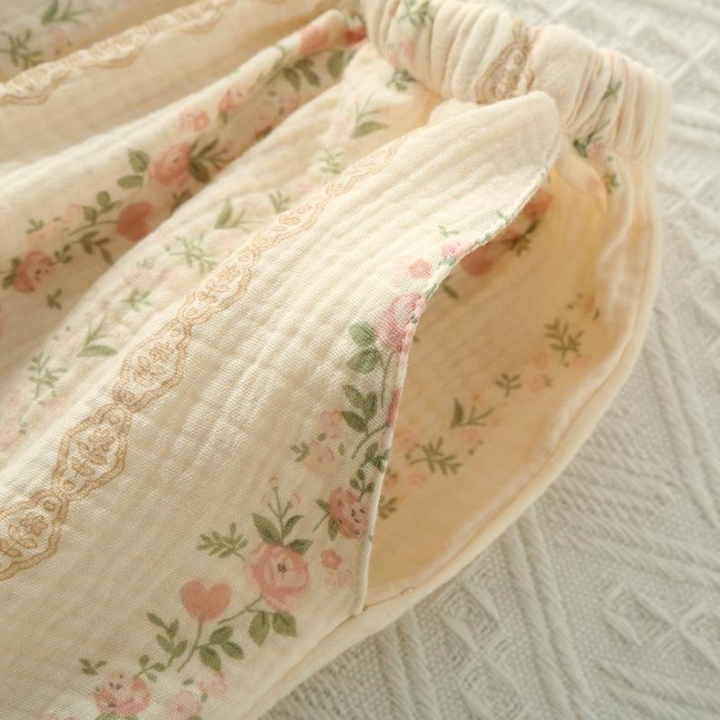 May include: Close-up of cream-colored pajama pants with a floral pattern of pink roses and green leaves. The pants have a drawstring waistband and a decorative lace trim. The pocket is lined with the same floral fabric.