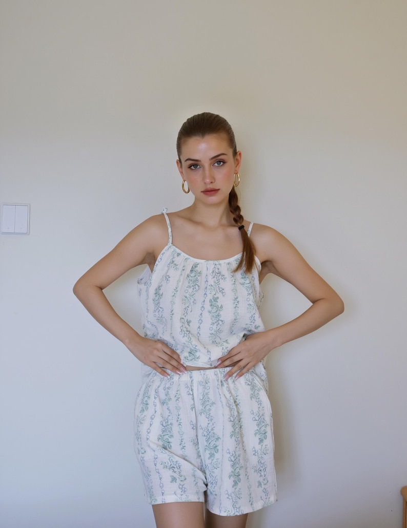 May include: A white pajama set with a green floral pattern. The set includes a camisole top with thin straps and matching shorts. The fabric has vertical stripes. The model is wearing gold hoop earrings.