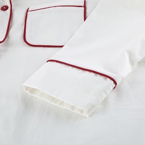 May include: Close-up of white pajama top with a red trim. The top features a pocket with red trim and a red button. The sleeve is visible, also trimmed in red. The fabric appears to be soft and comfortable.