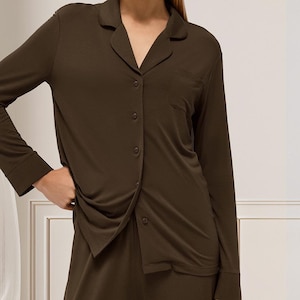 May include: Dark olive green pyjama set. The long-sleeved button-down shirt features a notched collar and a chest pocket. Matching bottoms are partially visible. The fabric appears soft and comfortable.