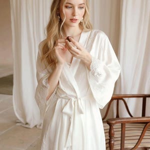 Könnte beinhalten: Ein weißes Satin-Kleid mit Spitzenbesatz, das mit einem passenden Bindegürtel in der Taille gebunden ist. Das Kleid hat weite Ärmel und wird über einem weißen Unterkleid getragen. Die Szene spielt in einem Raum mit transparenten Vorhängen und einem Holzstuhl.