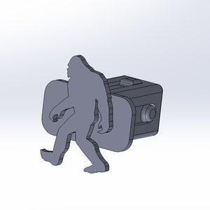 Pode incluir: Um chaveiro cinza impresso em 3D com uma silhueta de Pé Grande caminhando. O chaveiro está preso a um pequeno bloco de plástico cinza.