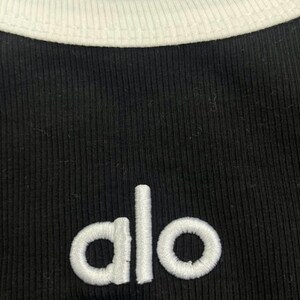 Może przedstawiać: Czarny prążkowany top z białym wykończeniem wok&oacute;ł dekoltu. Słowo "alo" jest wyhaftowane na biało z przodu. Top ma klasyczny, sportowy design.