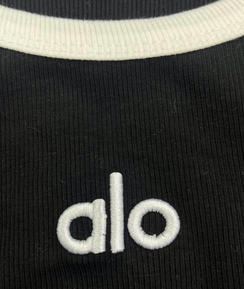 Może przedstawiać: Czarny prążkowany top z białym wykończeniem wok&oacute;ł dekoltu. Słowo "alo" jest wyhaftowane na biało z przodu. Top ma klasyczny, sportowy design.