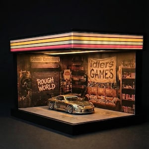 Op de afbeelding: Een zilveren model sportwagen wordt tentoongesteld in een diorama garage-omgeving. Het diorama heeft een gedetailleerd interieur met graffiti, een bank en planken. Het bord luidt "Rough World" en "Idlers Games". De bovenkant heeft kleurrijke strepen.
