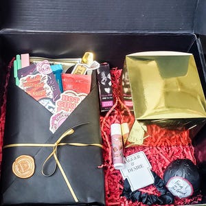 May include: A black gift box containing a black envelope sealed with a gold wax stamp, a gold box, lip balms, and a black scrunchie. The envelope holds stickers and a red and white item with text. The box is filled with red paper.