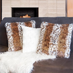May include: A collection of decorative throw pillows with faux fur covers. Two pillows feature a brown and white spotted pattern, while a third is solid white. A white faux fur throw is draped on the sofa.