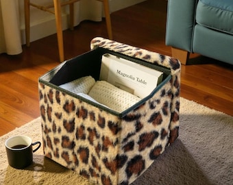 May include: A leopard print storage ottoman with a black interior, holding a book titled "Magnolia Table" and folded white blankets. A black mug with coffee sits on the rug next to the ottoman.