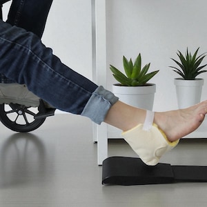 May include: A beige heel protector with a yellow interior is secured to the ankle with a white strap. A black support is positioned on the floor. The person is wearing blue jeans.