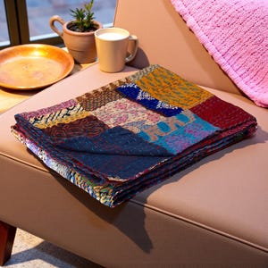 May include: A folded patchwork quilt with a variety of colors and patterns, including blue, red, yellow, and pink. The quilt is made of multiple fabric squares stitched together. The quilt is resting on a brown chair.