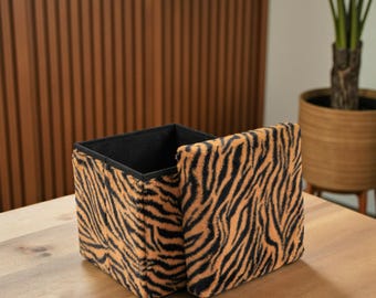 May include: A square storage ottoman with a tiger stripe pattern in orange and black. The lid is partially open, revealing a black interior. The ottoman is on a wooden surface.
