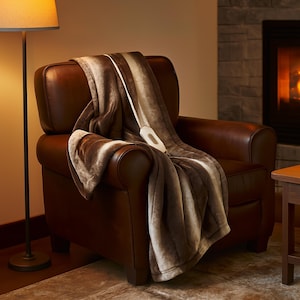 May include: A brown and beige heated blanket draped over a brown leather armchair. The blanket has a soft, plush texture and a white control panel. A floor lamp and a fireplace are visible in the background.