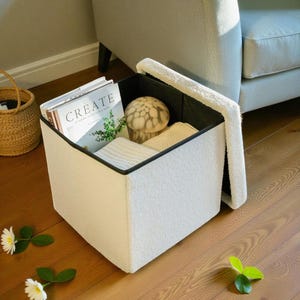 May include: A white storage ottoman with a black trim, open to reveal a magazine with the word "CREATE" on the cover, a decorative glass mushroom, and folded white towels. The ottoman is next to a light gray sofa.