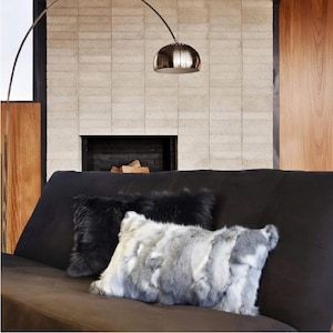 May include: A modern living room scene featuring a black sofa adorned with two faux fur pillows. One pillow is black, and the other is a mix of gray and white. A silver arc lamp hangs above the sofa, and a fireplace is in the background.