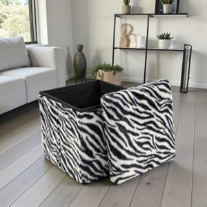 Könnte beinhalten: Quadratische Aufbewahrungshocker mit Zebra-Print-Design in Schwarz und Weiß. Der Deckel ist teilweise geöffnet und gibt den Blick auf das schwarze Innere frei. Der Hocker besteht aus einem weichen, strukturierten Material.