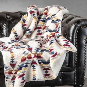 May include: A cream-colored sherpa blanket with a geometric pattern in red, blue, yellow, and gray. The blanket is draped over a black leather couch, showcasing its soft texture and design.