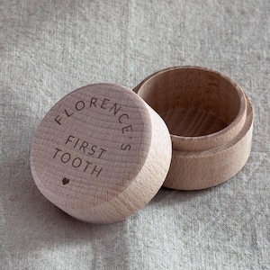 May include: A small, round, light-coloured wooden box with a removable lid. The lid is engraved with the text "FLORENCE'S FIRST TOOTH" and a small heart symbol. The box has a smooth, natural wood grain finish and a simple, cylindrical shape.