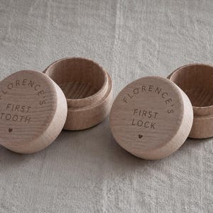 May include: Two round, light-coloured wooden keepsake boxes with removable lids. One lid is engraved with the text FLORENCE'S FIRST TOOTH and a small heart, while the other lid is engraved with FLORENCE'S FIRST LOCK and a small heart.
