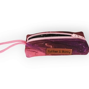 Könnte beinhalten: Eine kleine rosa Reißverschlusstasche mit einem rosa Riemen und einem braunen Lederanhänger mit der Aufschrift "Toffee & Bomby". Die Tasche hat ein rosa-violettes Marmormuster.