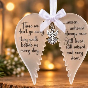 May include: A white heart-shaped ornament with angel wing cutouts, featuring black text that reads: Those we don't go away, they walk beside us every day... unseen, unheard, always near. Still loved, still missed and very dear. A white ribbon bow and silver angel charm.