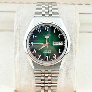 Op de afbeelding: Een zilveren Seiko 5 automatisch horloge met een groene wijzerplaat en Arabische cijfers. Het horloge heeft een roestvrijstalen band en een dag-datumweergave. De wijzerplaat leest "Automatic 17 Jewels" en "Made in Japan".