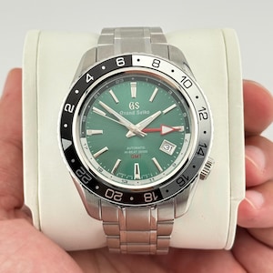 May include: A stainless steel Grand Seiko watch with a green dial and a black and silver bezel. The watch features a date display and a silver band. The watch is held in a person's hand.