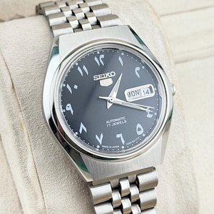 Op de afbeelding: Een zilverkleurig Seiko-horloge met een zwarte wijzerplaat en Arabische cijfers. Het horloge heeft een dag- en datumweergave, een roestvrijstalen band en de tekst "AUTOMATIC 17 JEWELS". Het is gemarkeerd met "MADE IN JAPAN".