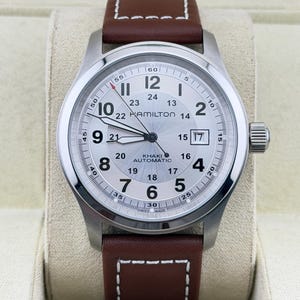 May include: A Hamilton Khaki Automatic wristwatch with a silver dial, black Arabic numerals, and a 24-hour inner track. The watch features a date window at the three o'clock position, luminous hands, a red-tipped second hand, and a brown leather strap.