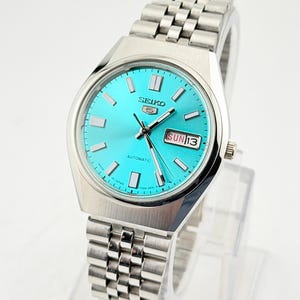 May include: A silver-toned Seiko 5 automatic watch with a vibrant turquoise dial. The watch features a stainless steel band, rectangular hour markers, and a day-date display. The watch face displays "SUN 13".