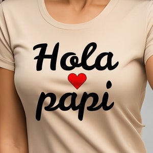May include: A beige t-shirt with the text "Hola papi" in black with a red heart between the words.