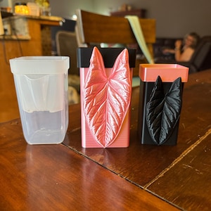 May include: Three square planters arranged in a row. The first is clear plastic. The second is a pink planter with a metallic pink leaf relief. The third is a black planter with a black leaf relief and a pink rim at the top.