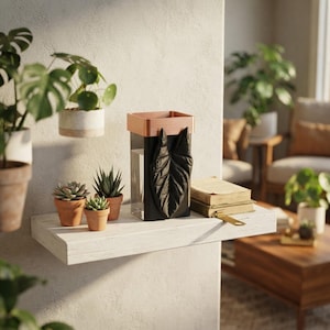May include: A rectangular vase with a black base and a copper-colored rim, featuring a prominent black leaf-shaped relief on the front. The vase is positioned on a white shelf alongside three small potted succulents and two stacked books.