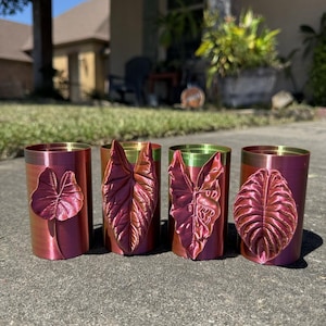 May include: Four cylindrical cups with a metallic finish that shifts between red, pink, and green. Each cup features a distinct, raised, three-dimensional leaf design on the front, showcasing intricate vein patterns and a glossy, iridescent texture.