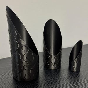 May include: Three matte black cylindrical scoops of varying heights, each featuring a raised, repeating organic pattern on the exterior surface. The scoops have an angled, open top edge and a flat base, arranged in a row from largest to smallest.