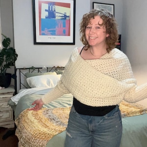 May include: A woman wearing a white crocheted sweater with a cowl neck and blue jeans. She is standing in front of a bed with a green bedspread and a yellow and white crocheted throw blanket.