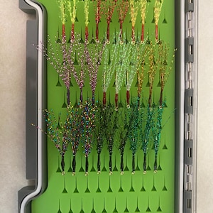 May include: A green fly fishing box with a variety of colorful flies arranged in rows. The flies are made with different materials, including feathers, yarn, and beads.