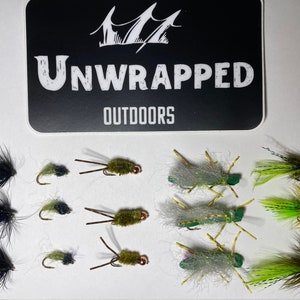 May include: A collection of 12 fly fishing lures, including black and green, green and white, and brown and green flies. The flies are arranged in three rows of four. The image also includes a black and white sticker with the text "Unwrapped Outdoors".