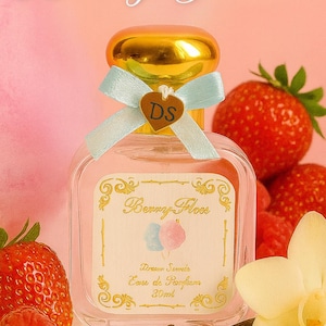 Puede incluir: Un frasco de perfume con tapa dorada y lazo azul claro, etiquetado como "Berry Floss". El frasco está rodeado de fresas, frambuesas y una vaina de vainilla. El fondo es de color rosa suave.